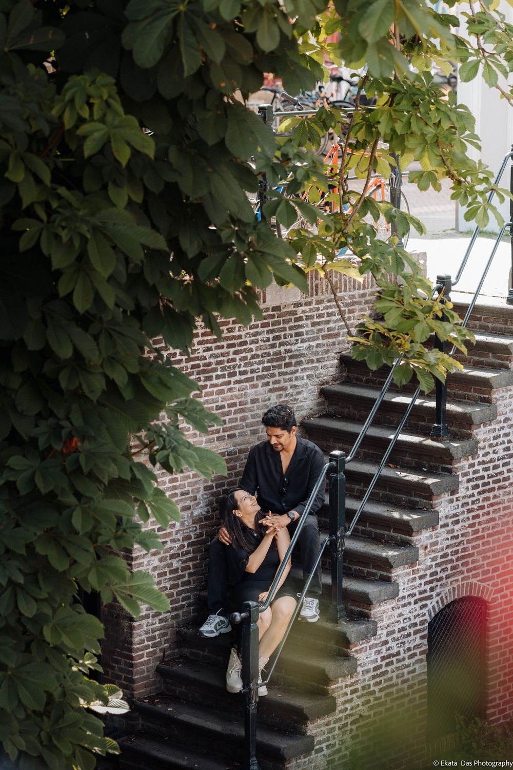Couple sitting on brick staircase surrounded by lush green foliage, sharing a romantic moment outdoors.