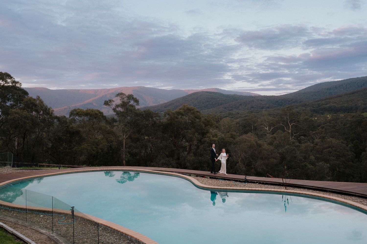 Couple standing by an infinity pool with stunning mountain views and dramatic cloudy skies in a lush Australian landscape.