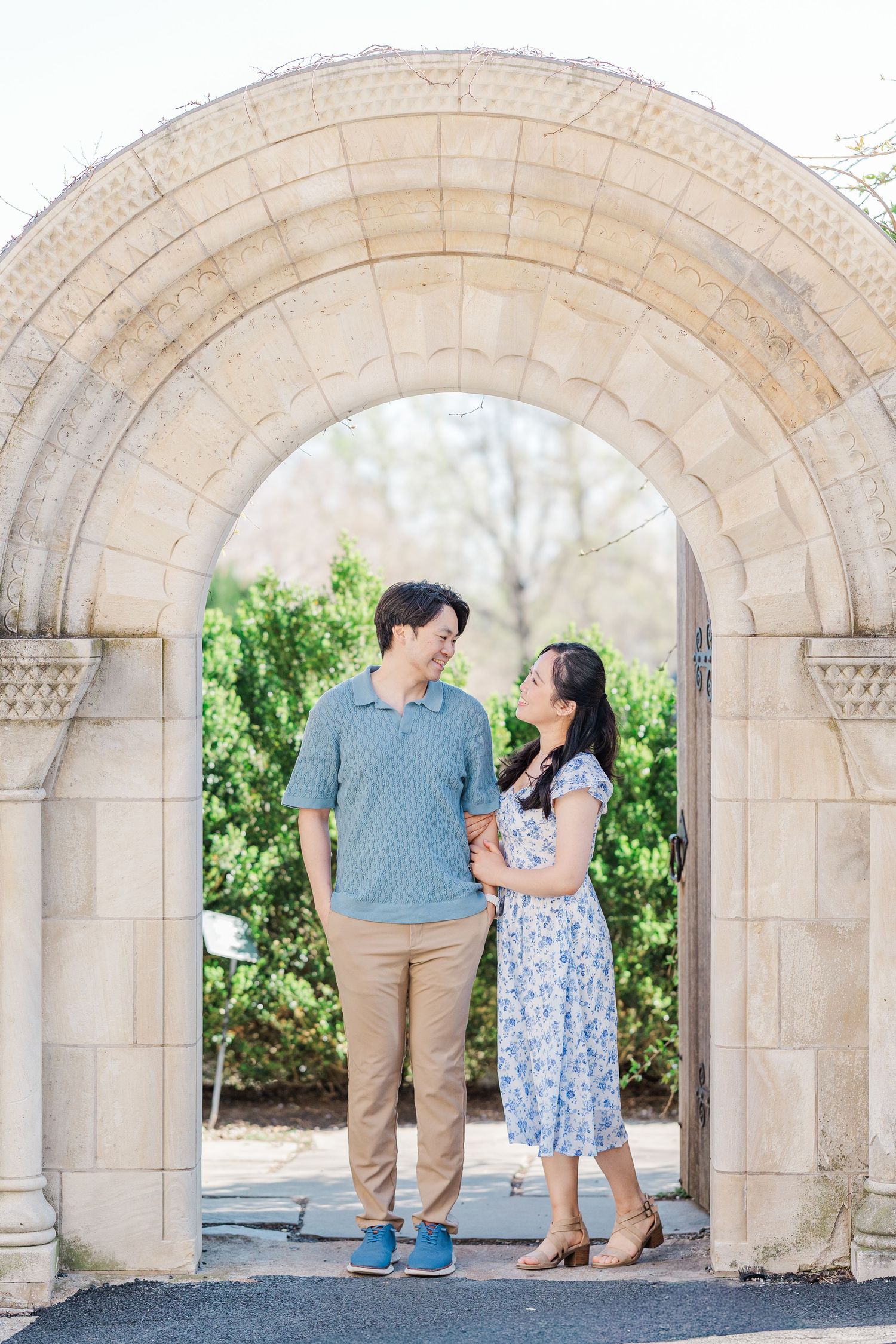 Couple standing together under a stone archway in a garden, smiling at each other in casual spring attire.