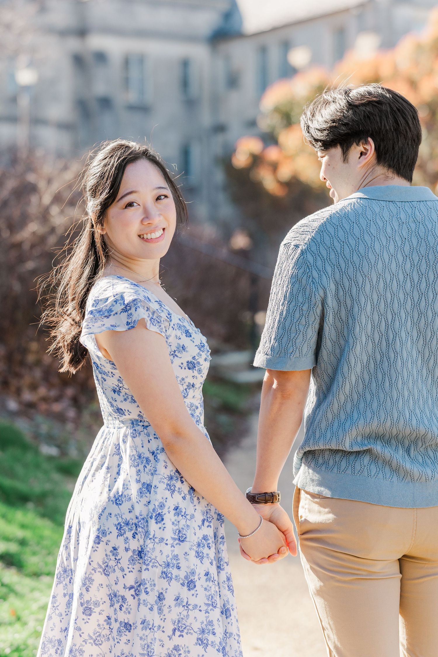 A smiling woman in a floral dress holds hands with a man outdoors on a sunny day.