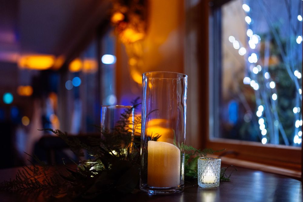 Cozy candlelit table with glass cylinder candles and holiday lights glowing through a dark window.