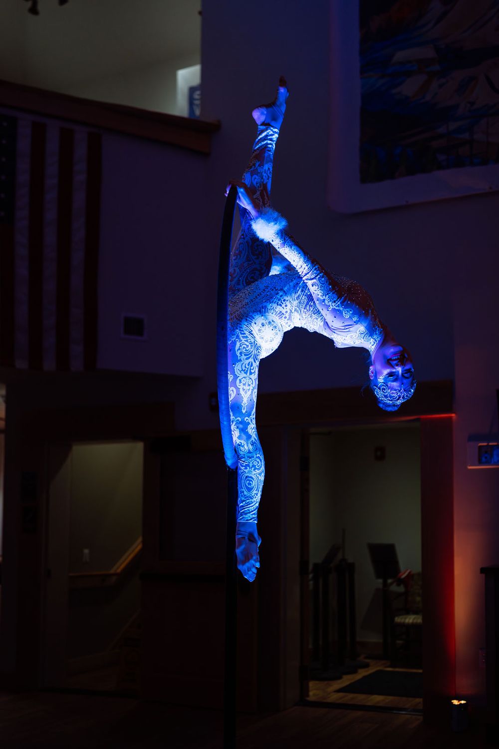 Acrobat performing a vertical pole split under dramatic blue UV lighting in a darkened indoor venue.