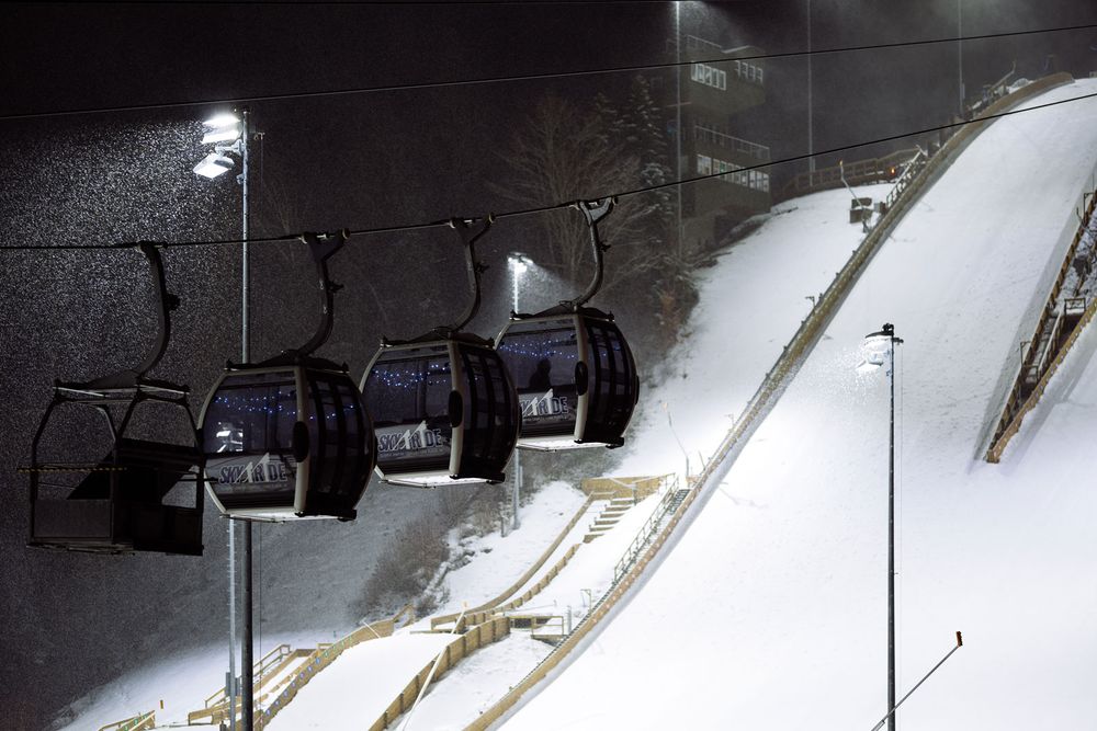 Night ski lift chairs hang over a snow-covered slope illuminated by bright stadium lights at a winter resort.