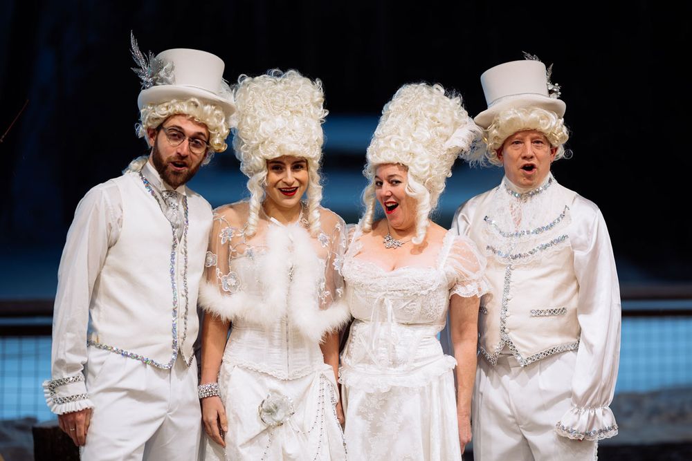 Four performers in ornate white baroque costumes and tall powdered wigs pose together on a theatrical stage.