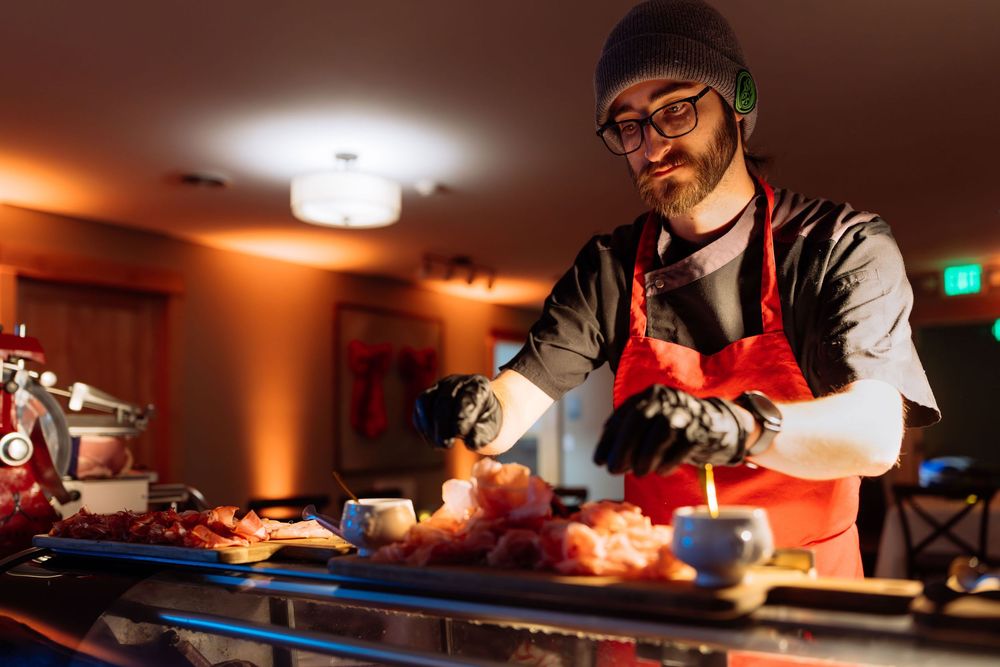 Bearded chef in red apron and black gloves preparing charcuterie at a dimly lit food station.