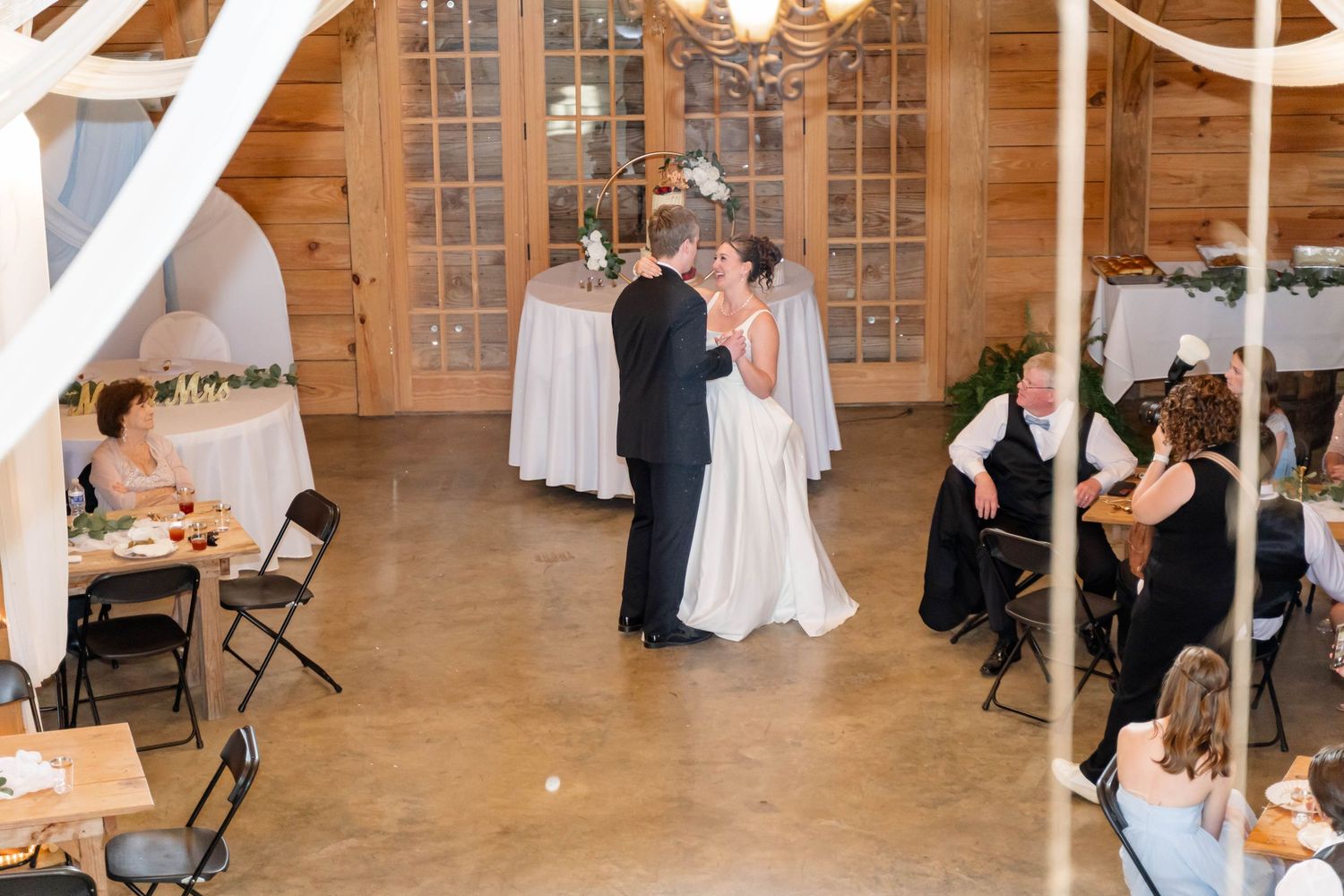 Bride and groom share first dance in rustic wooden barn wedding venue surrounded by seated guests at decorated tables.