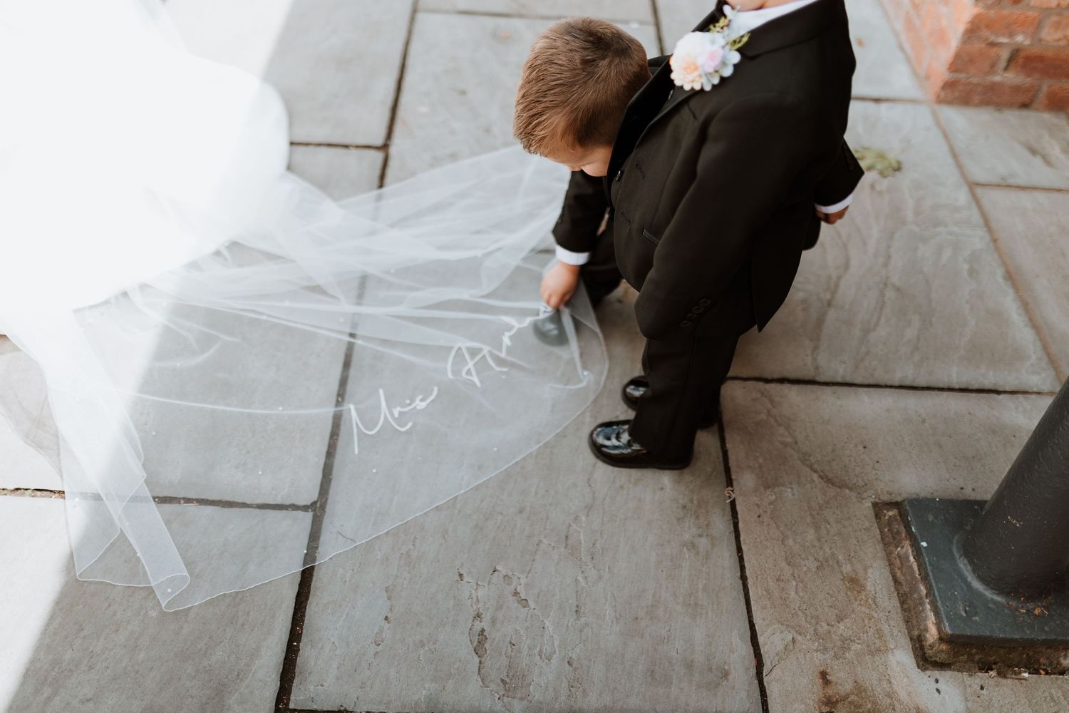 A young ring bearer in a black suit touches a bridal veil with Mrs. written on it on stone pavement.