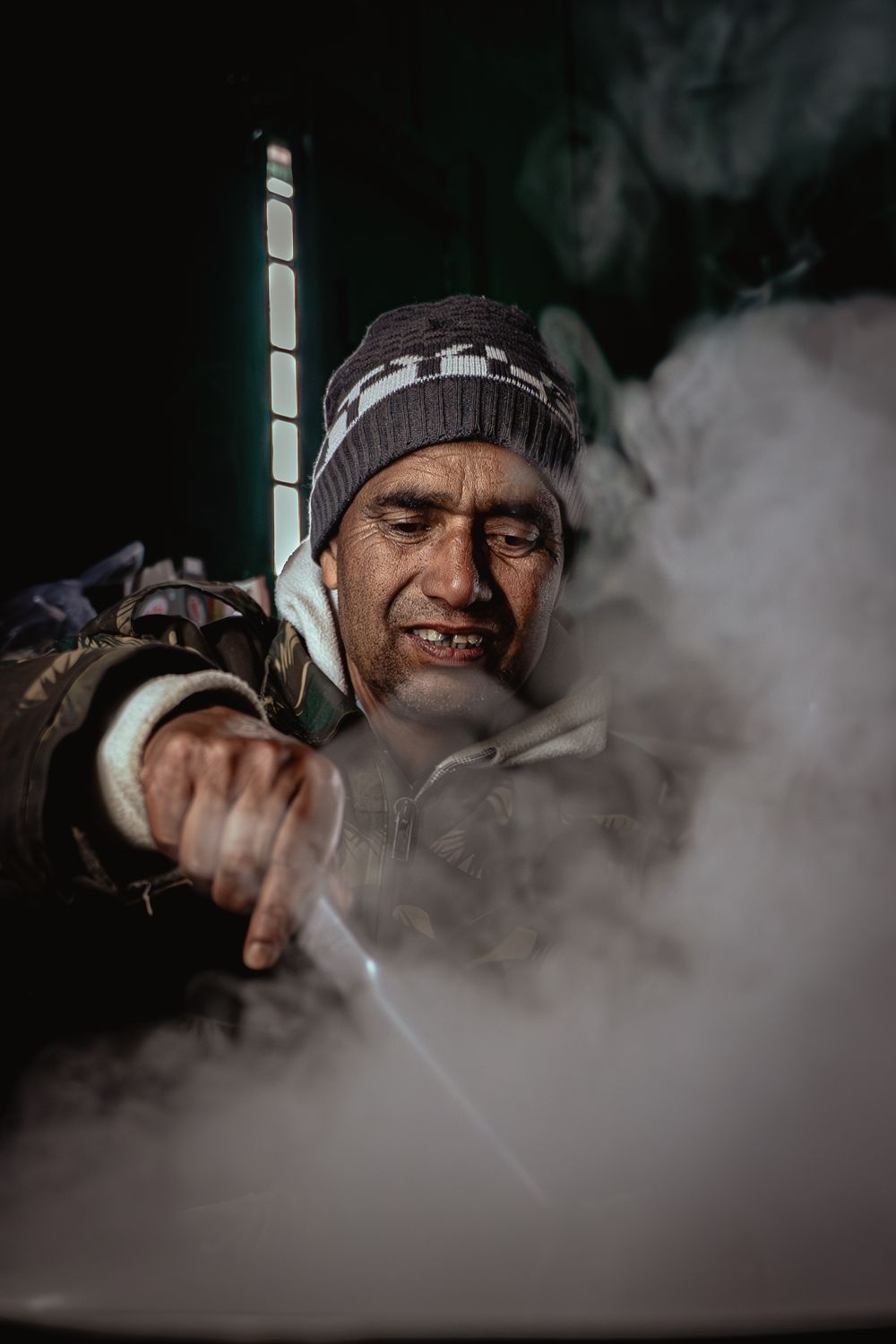 A weathered man in a knit cap leans over a steaming pot in a dark setting, smoke billowing around him.