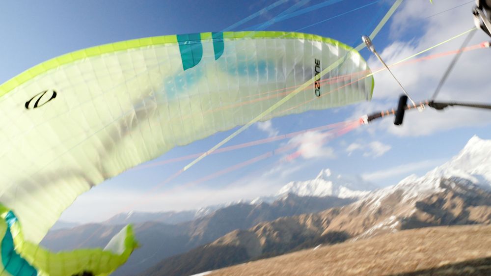 Colorful paraglider canopy soaring over snow-capped mountain peaks under a clear blue sky.