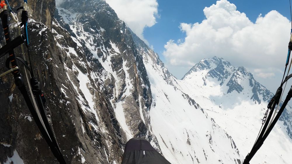 Aerial view of a steep snow-covered mountain ridge with rocky cliffs and distant peaks under a cloudy sky.