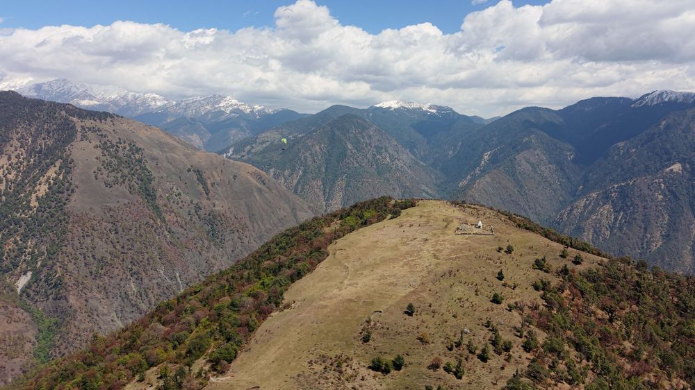 Aerial view of a lush mountain ridge with sweeping valley views and snow-capped peaks under a partly cloudy sky.