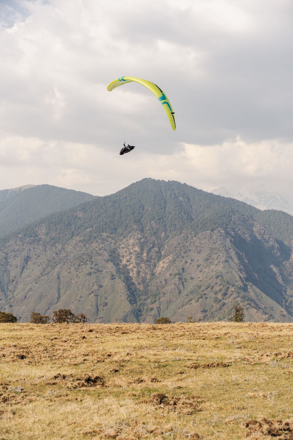 A paraglider with a yellow canopy soars over a vast mountain landscape under a cloudy sky.