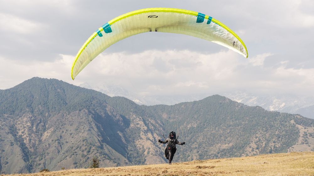 A paraglider soars above mountain terrain with a bright yellow-green canopy against a cloudy sky.