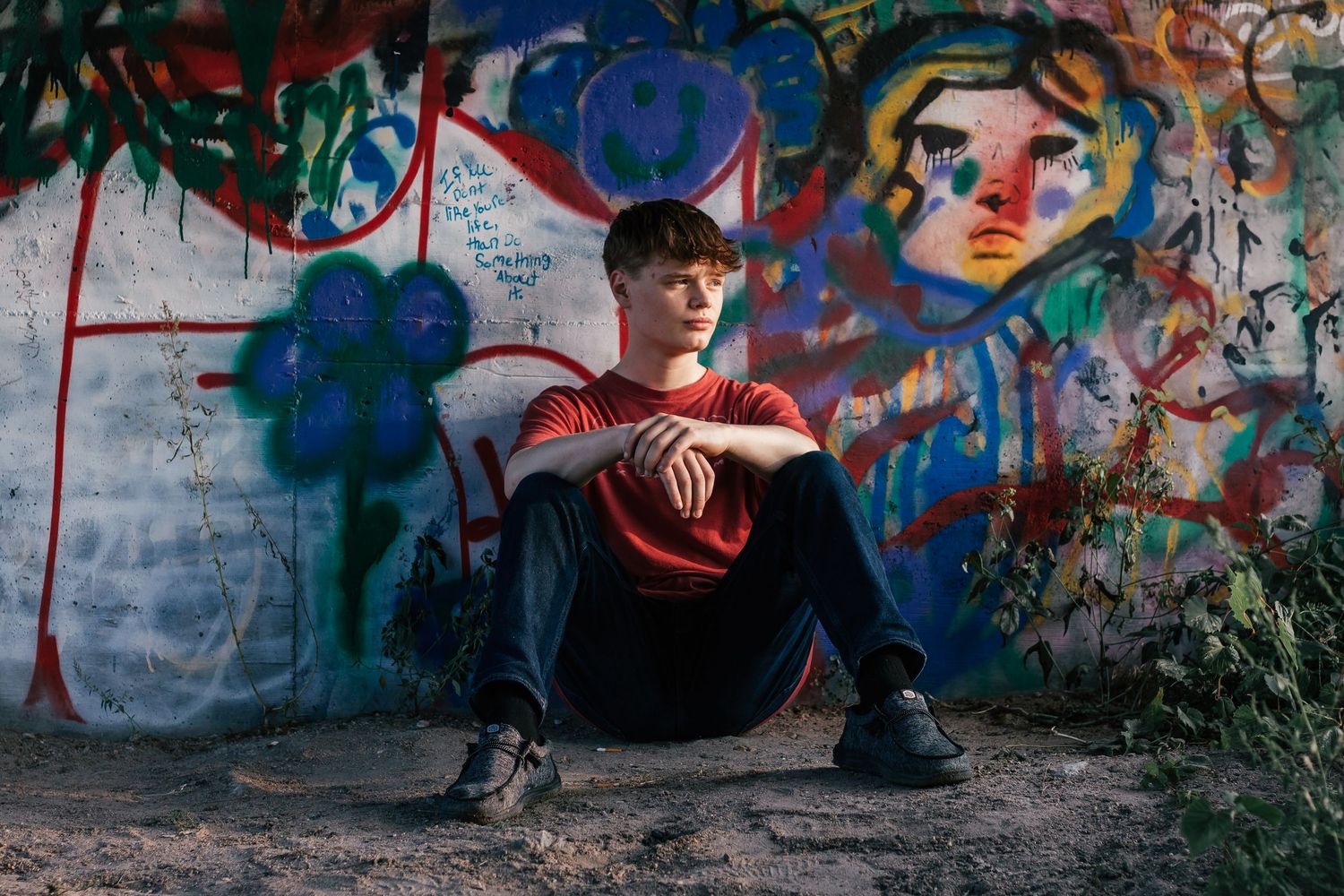Young person sitting against a colorful graffiti wall outdoors, wearing a red shirt and black pants.