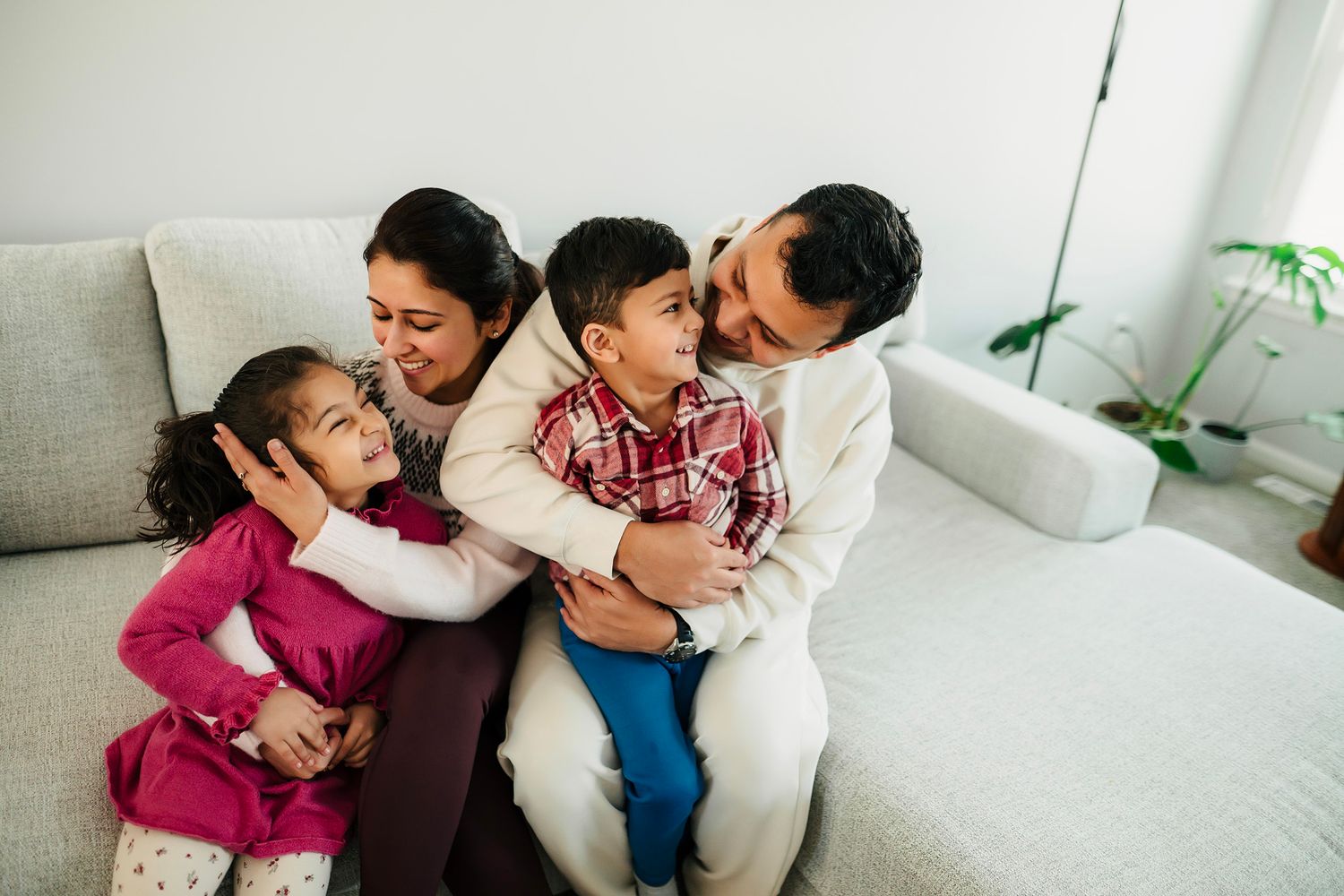 Happy family of four sitting together on a cozy white sofa, parents hugging two young children at home.