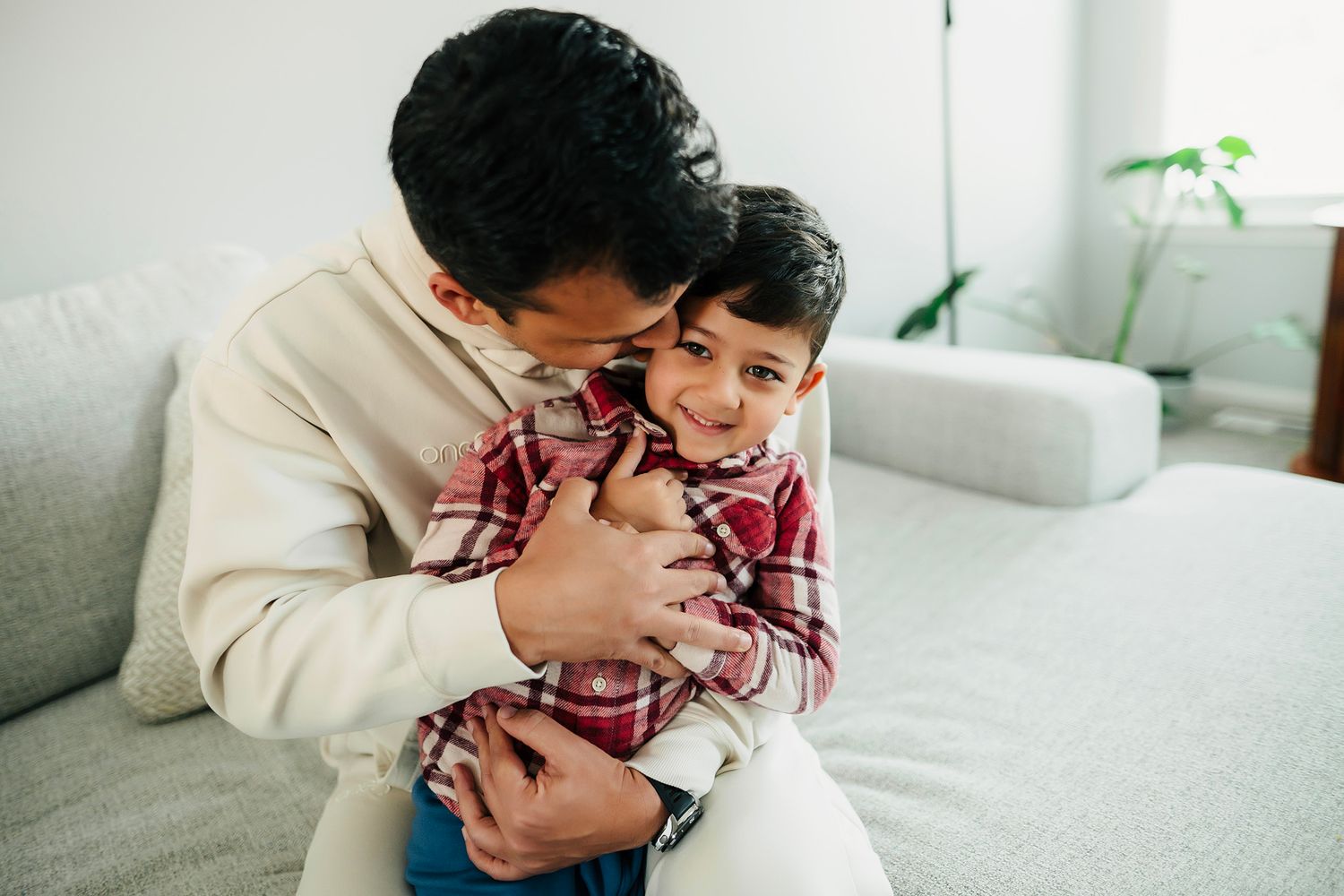 A father lovingly hugs his smiling young son on a cozy sofa at home.