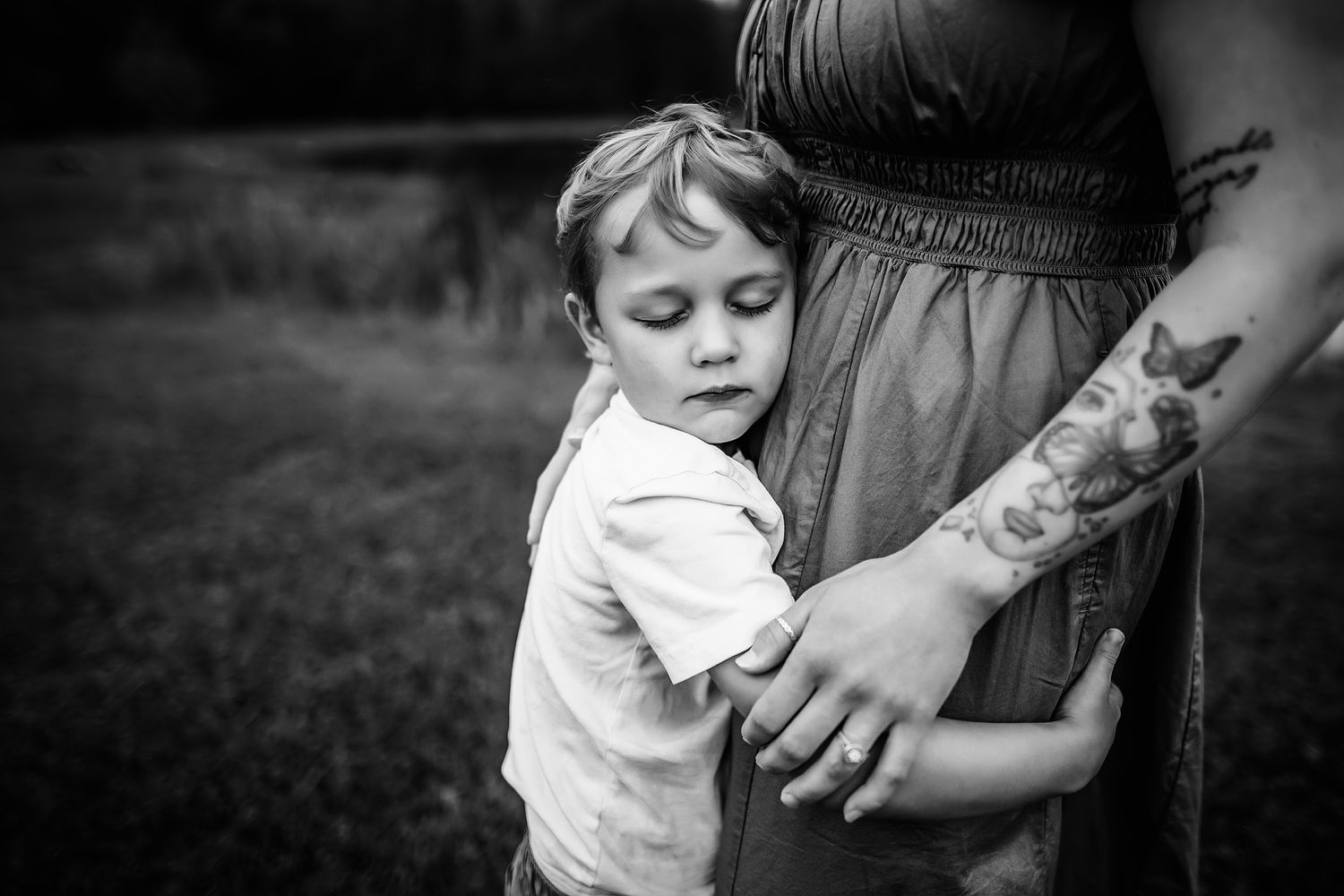 Black and white photo of a young boy hugging his pregnant mothers belly outdoors in a grassy field.