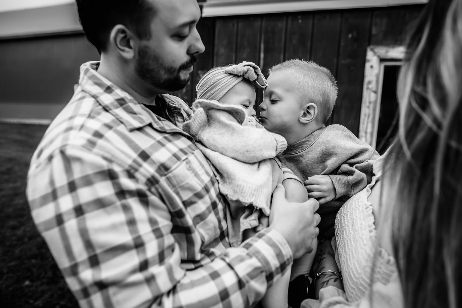 Black and white photo of a bearded father holding two babies who are kissing each other outdoors.