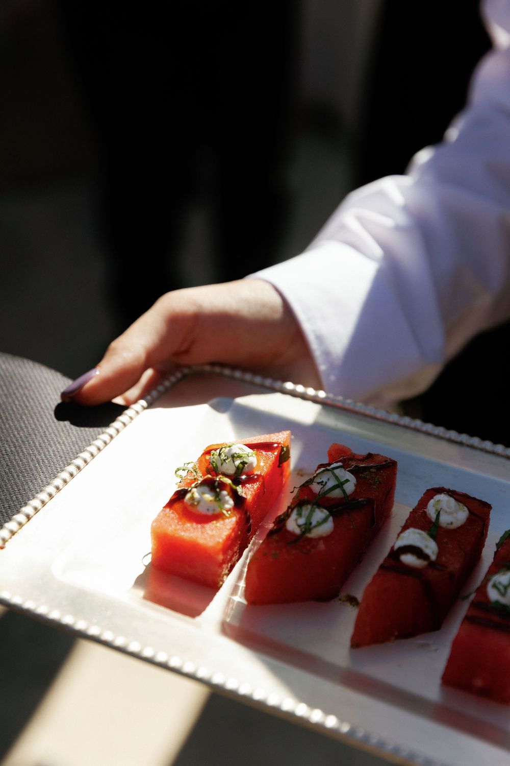 A chef in white uniform holds a silver tray with elegant watermelon appetizers topped with cream cheese and herbs.