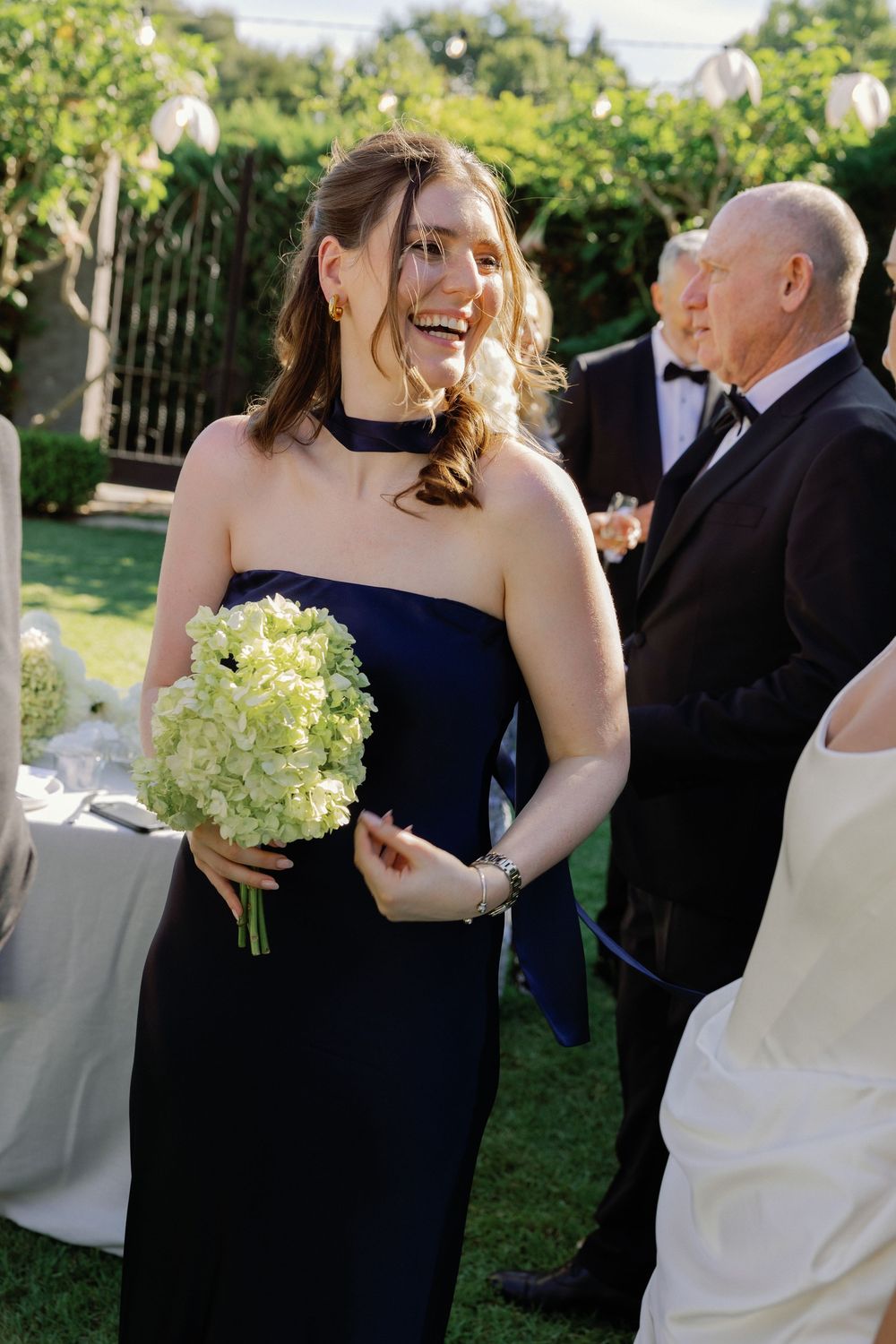 Smiling bridesmaid in navy blue strapless gown holding white hydrangea bouquet at outdoor wedding reception.