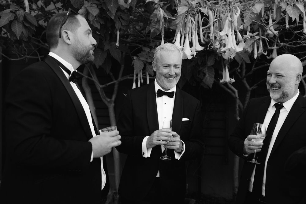 Three men in tuxedos laughing and holding champagne glasses outdoors in a black and white photo.