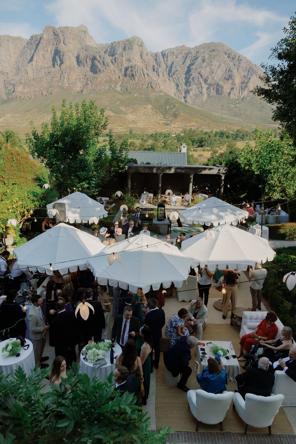 Outdoor wedding reception with white umbrellas, guests mingling on a patio, and stunning mountain views in the background.