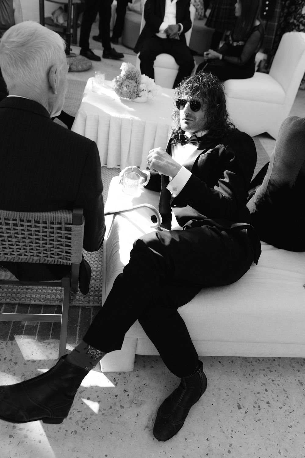 Black and white photo of a man in a tuxedo and sunglasses sitting on a white sofa at a formal event.