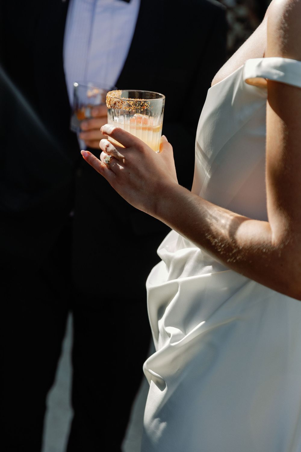 Bride in white off-shoulder gown holds a cocktail glass with gold rim at a wedding reception.