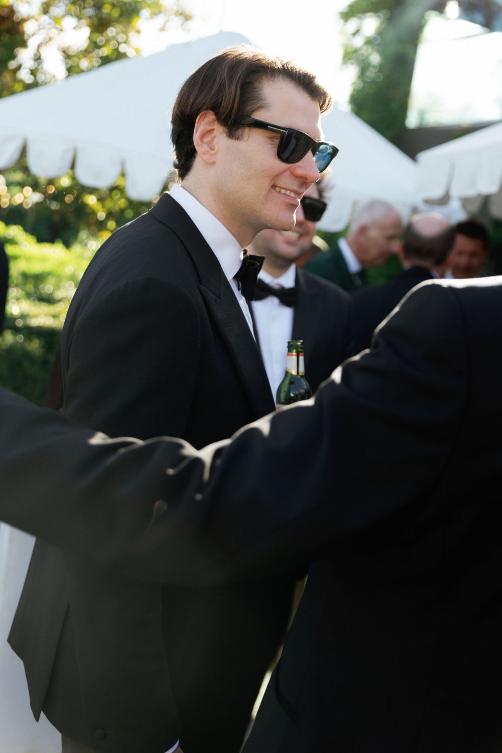 Stylish groom in black tuxedo and sunglasses smiles at outdoor wedding reception holding a bottle.