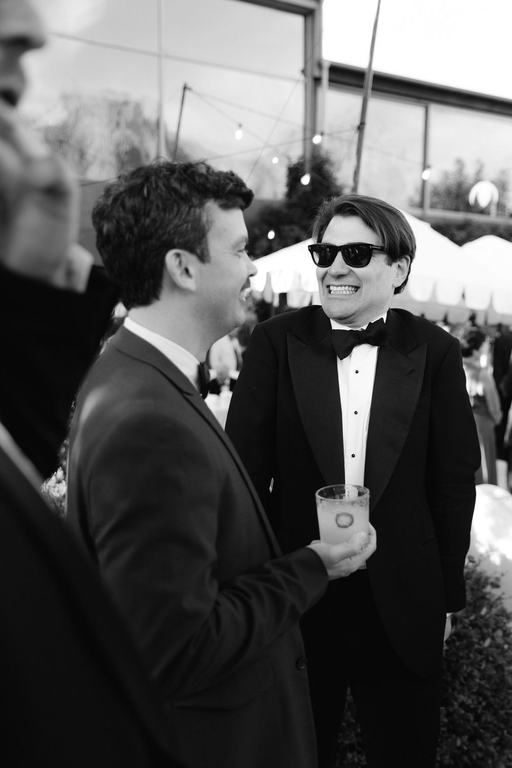Two men in tuxedos chat at an outdoor formal event, one wearing sunglasses and holding a drink, in black and white.