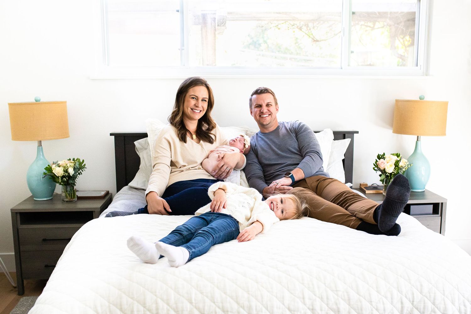 Happy family of four with two young children smiling together on a white bed in a bright, cozy bedroom.