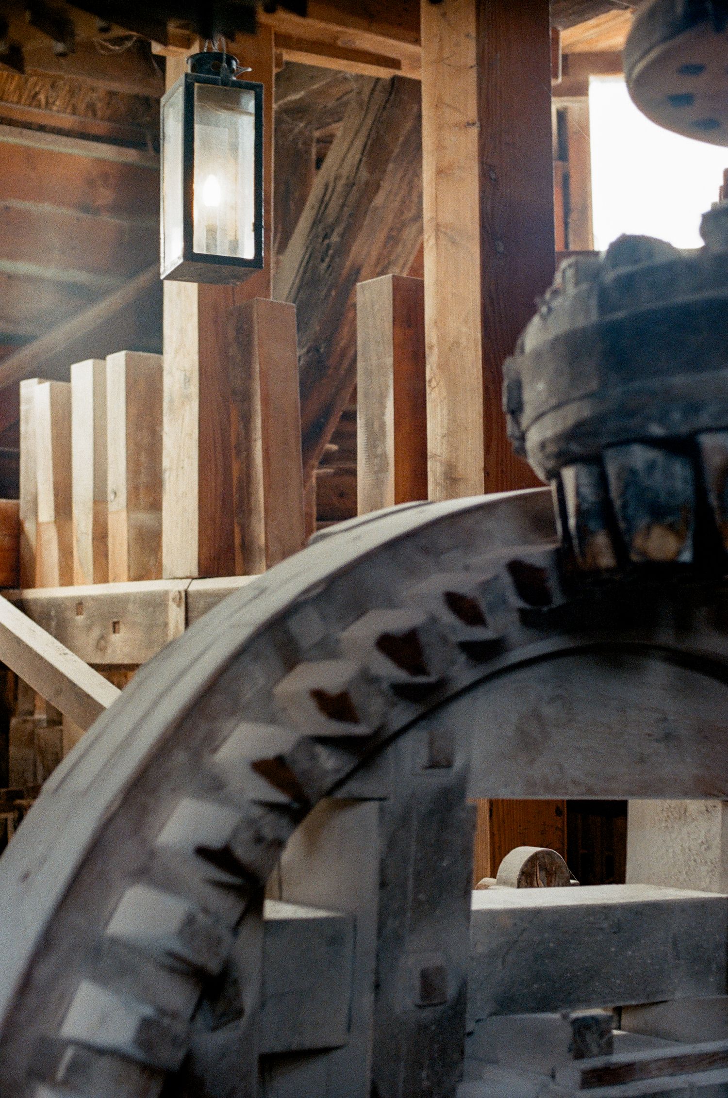 Large metal gear wheel inside a rustic wooden mill building with a vintage lantern light glowing in the background.