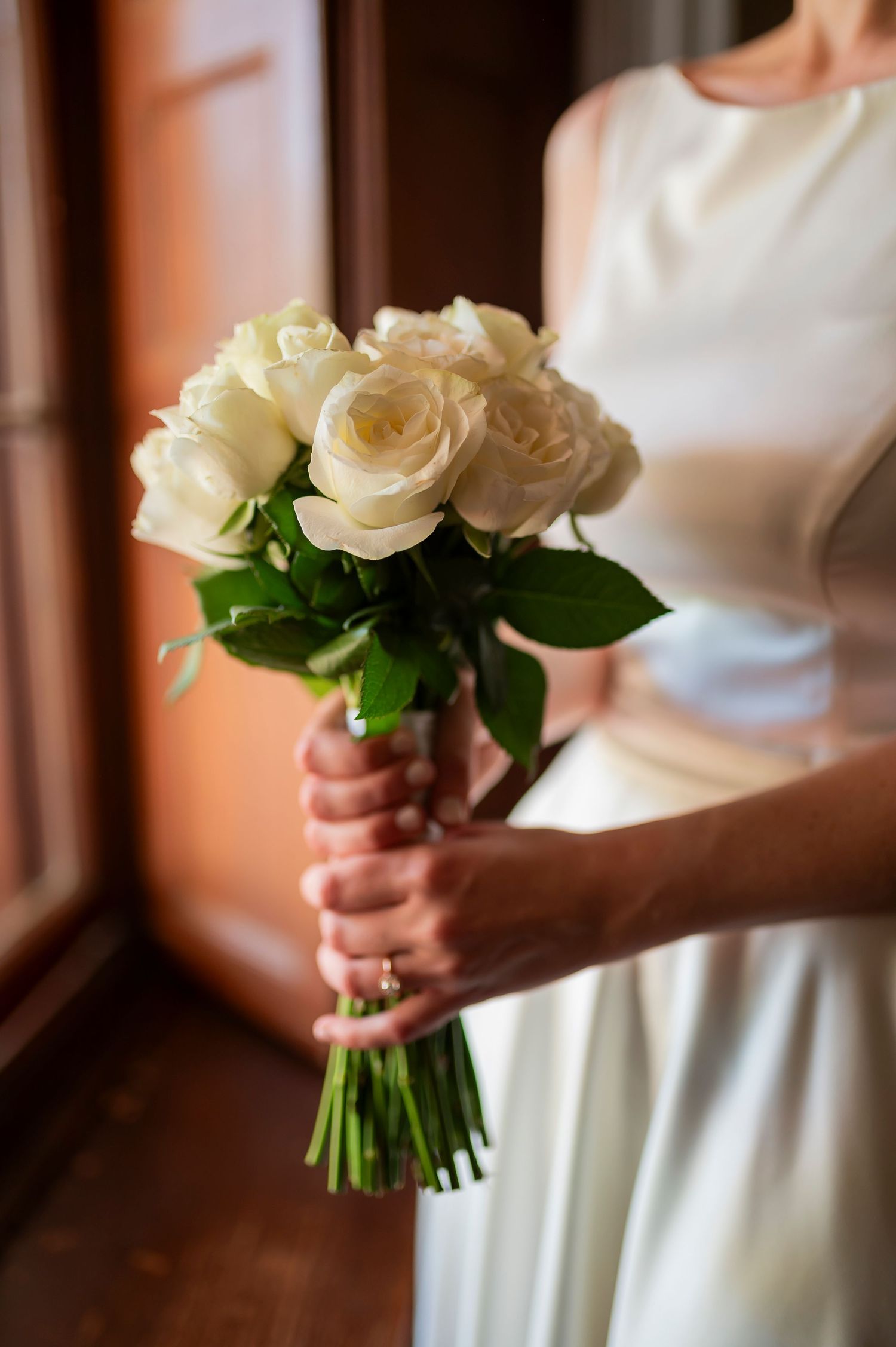Bride in white dress holding elegant bouquet of white roses with green stems at wedding ceremony.