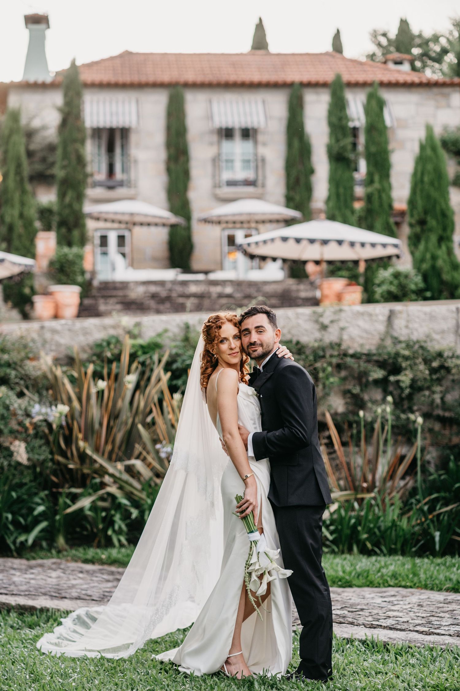 Bride and groom embrace in garden of elegant stone villa with cypress trees and terracotta roof.