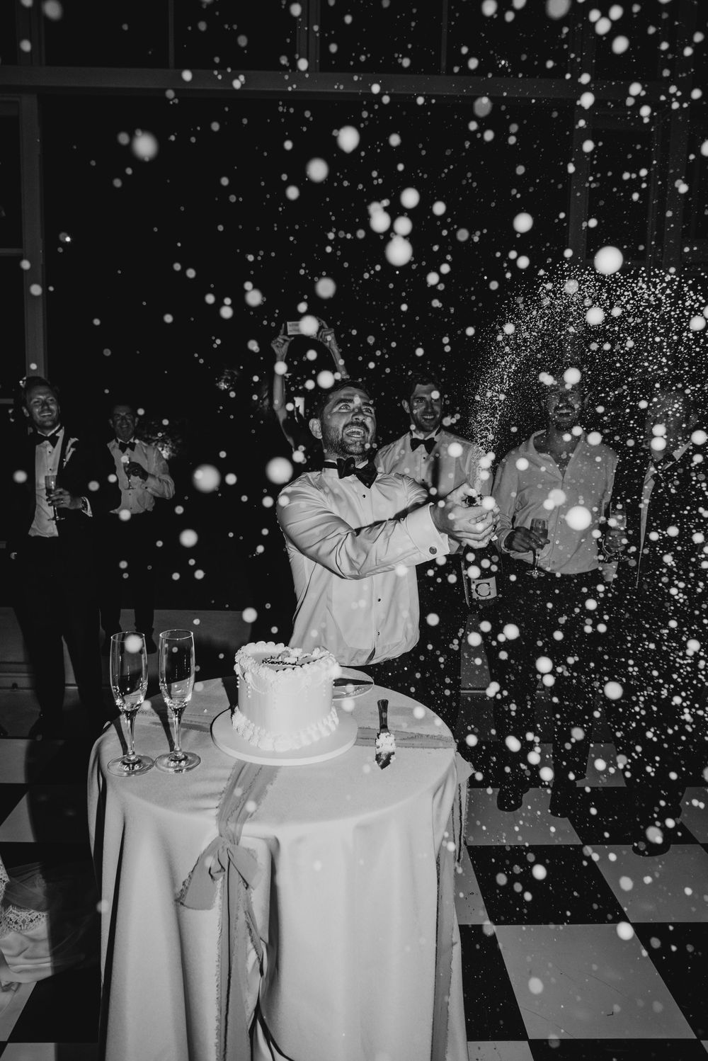 Groom pops champagne over wedding cake on checkered dance floor, surrounded by groomsmen in black and white photo.