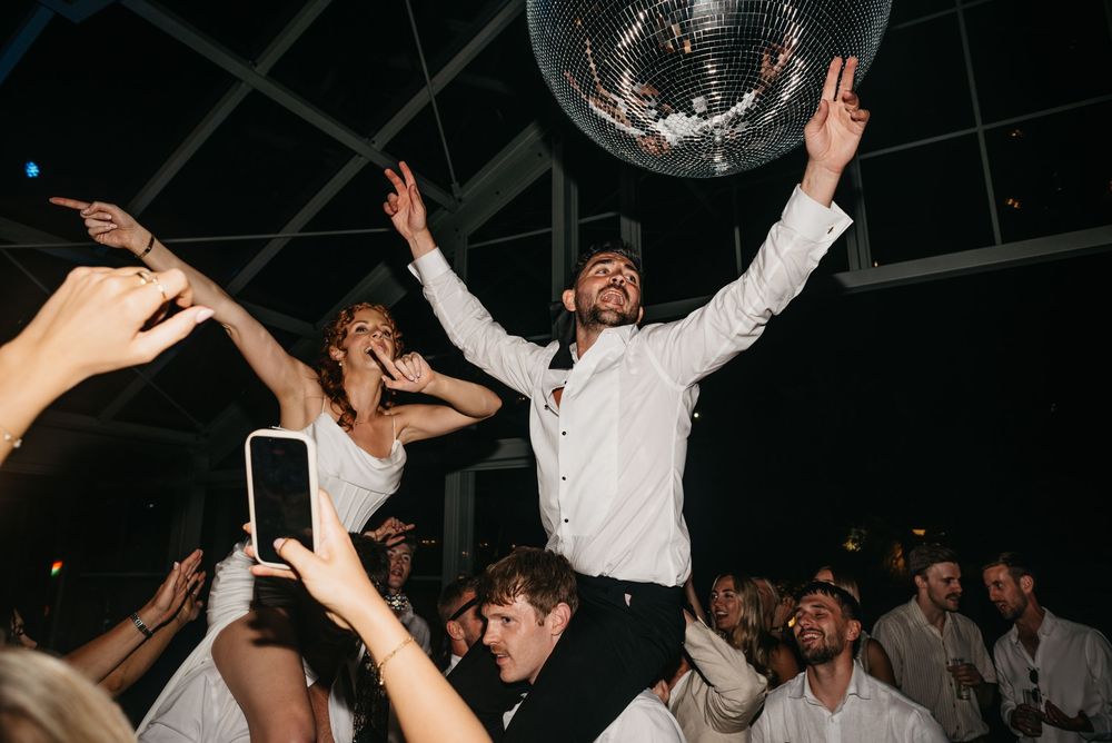 Bride and groom celebrate on dance floor under disco ball with raised arms at lively wedding reception.