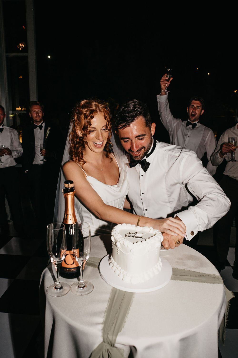 Bride and groom cutting white wedding cake beside champagne bottle and glasses at elegant evening reception.