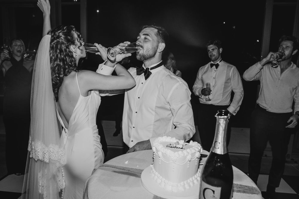 Black and white photo of bride and groom drinking champagne together at wedding cake table.