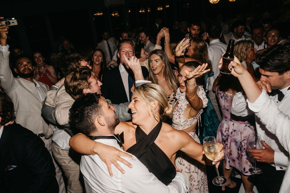 Joyful wedding reception dance floor with bride in black halter dress, groom, and celebrating guests at night.