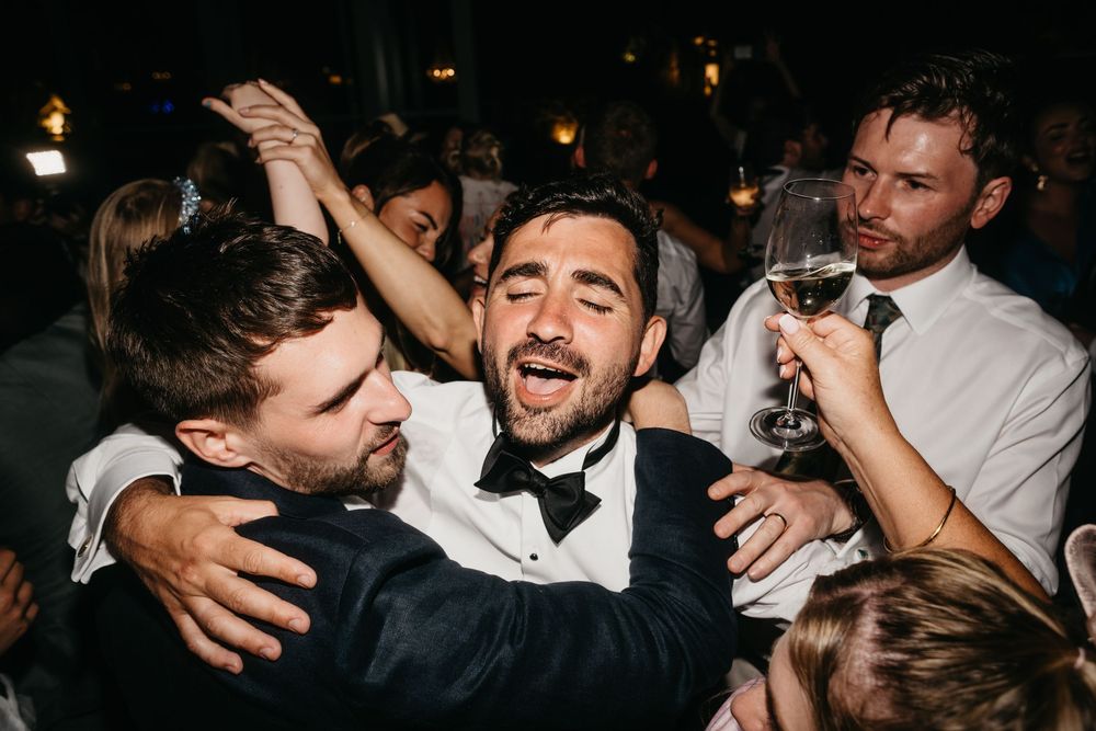 Joyful wedding reception guests celebrating and dancing together, man in bow tie laughing with friends holding champagne.