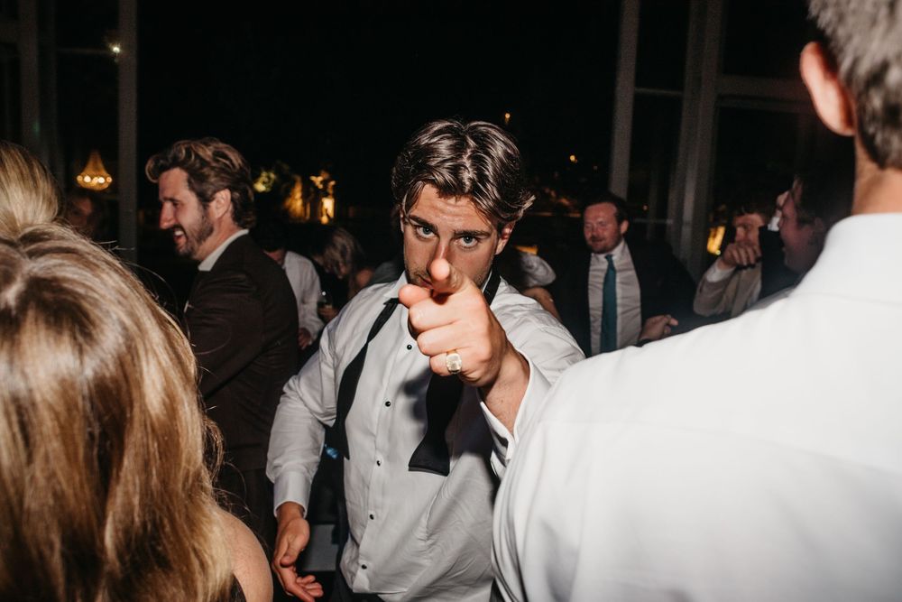 Man in white dress shirt pointing at camera at a lively evening wedding reception dance floor.