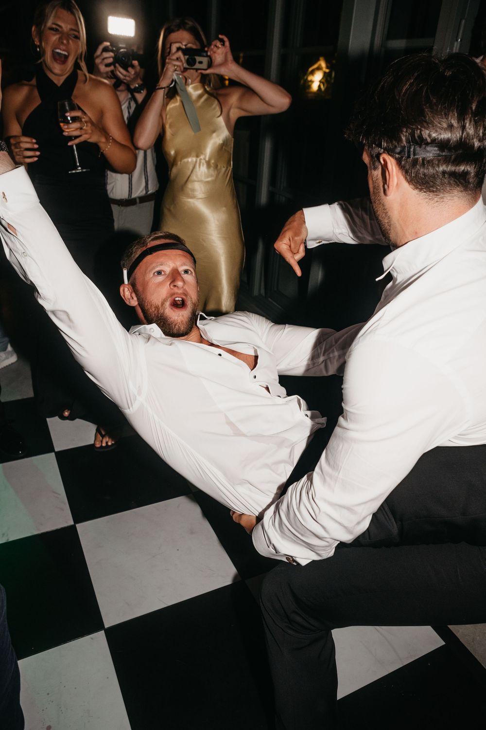 Two men dancing energetically on a checkered dance floor at a party while guests laugh and photograph them.