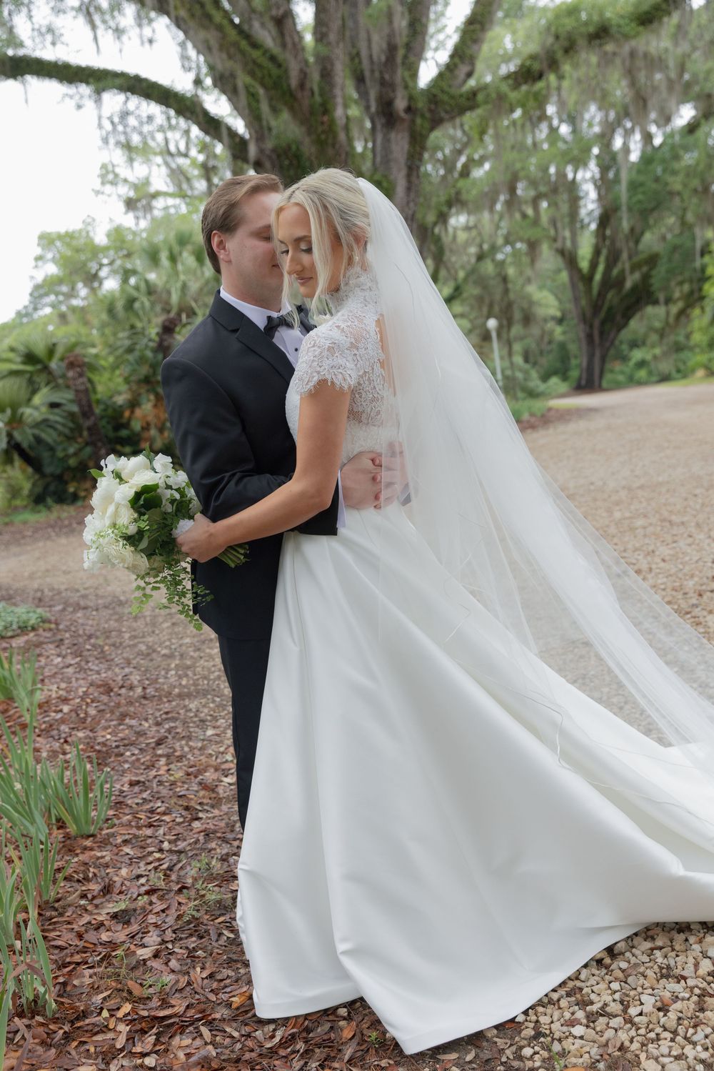 Bride and groom embrace outdoors under moss-draped oak trees, bride wearing lace gown with flowing veil holding white bouquet.