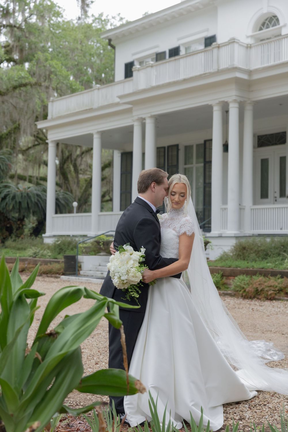 Bride and groom embrace in front of a white Southern mansion, bride holding white hydrangea bouquet with flowing veil.