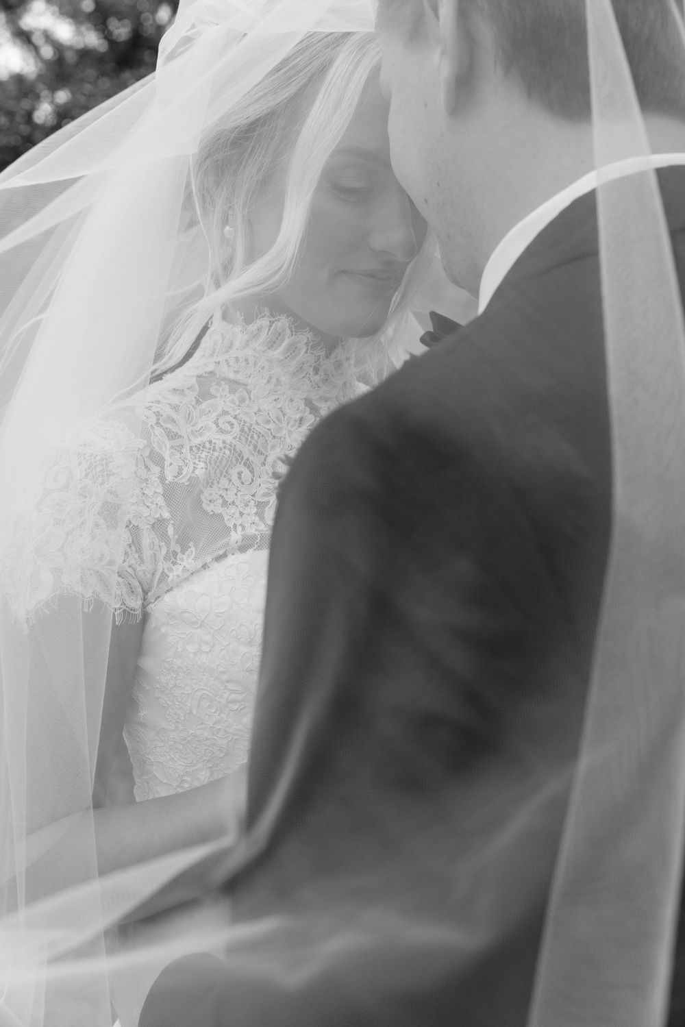 Black and white wedding photo of bride in lace gown and groom embracing under flowing veil.