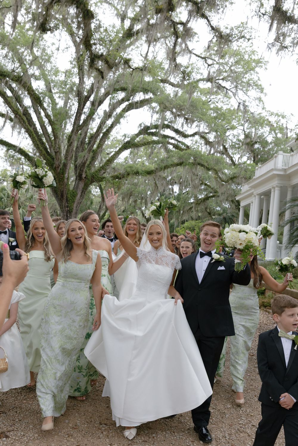 Joyful wedding party celebrates outdoors under Spanish moss oak trees, bride in white gown with groom in tuxedo.
