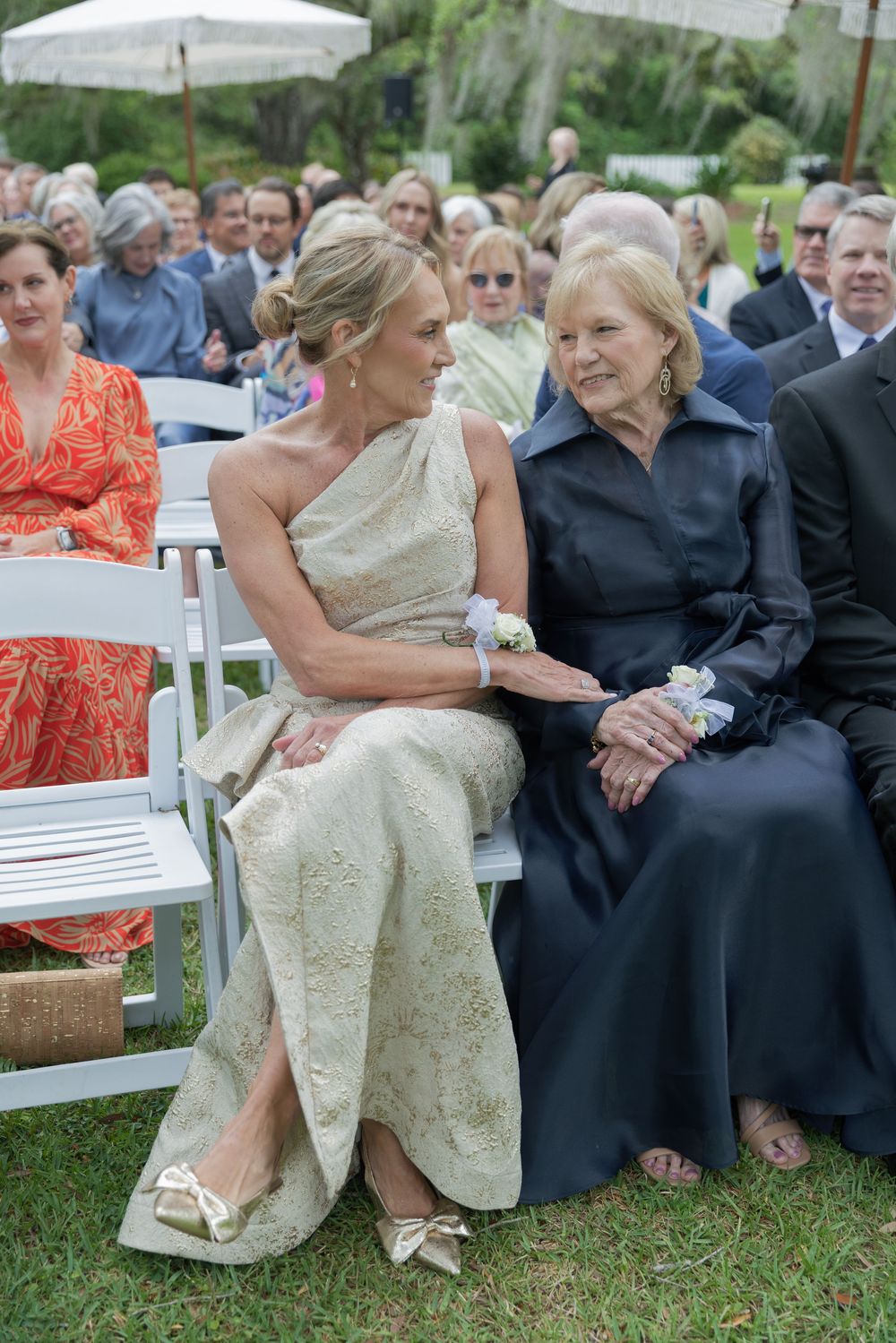 Two elegantly dressed women share a tender moment seated at an outdoor wedding ceremony on a sunny day.