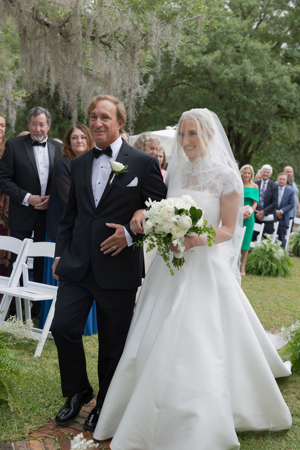 Bride in white lace gown walks down outdoor aisle with escort in tuxedo, guests seated on white chairs.