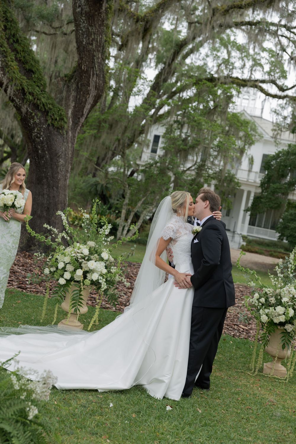 Bride and groom share first kiss at outdoor wedding ceremony surrounded by white floral arrangements and Spanish moss trees.