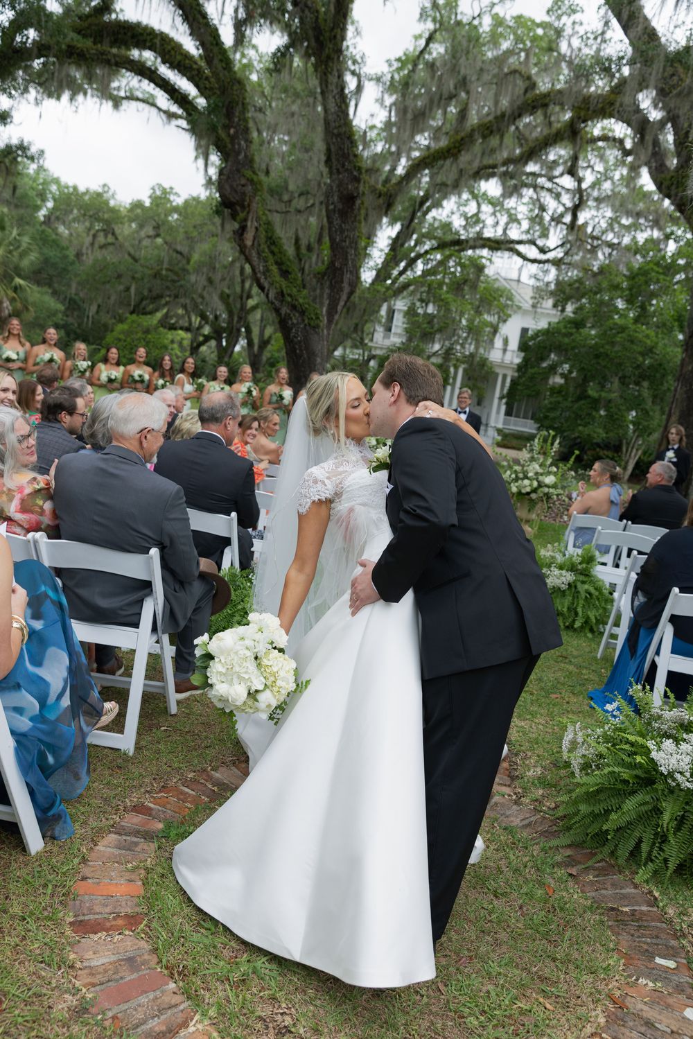 Bride and groom share a first kiss at outdoor wedding ceremony with guests seated in white chairs under oak trees.